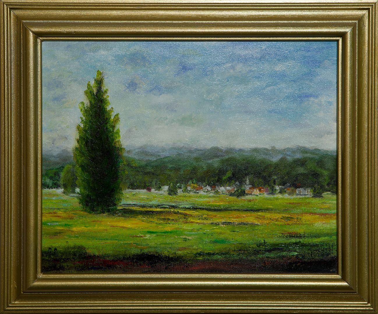 A tall, dark evergreen stands alone in a wide green meadow, with a small village of red-roofed houses and wooded hills beyond under a cloudy sky.